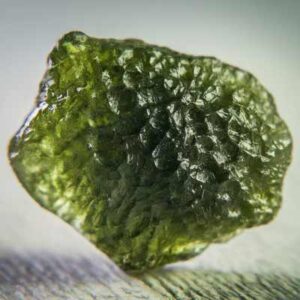 A close-up image of a raw Moldavite specimen, highlighting its textured surface, translucent green color, and irregular, otherworldly shape.