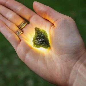 A close-up photo of a person's hand holding a Moldavite crystal over their heart chakra, bathed in a soft green light.