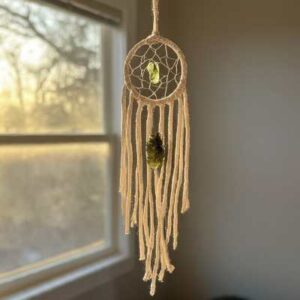 A dreamcatcher with a Moldavite crystal woven into the center, hanging above a bed with soft light filtering through the window.