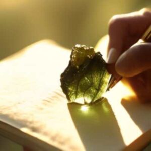 A close-up photo of a person holding a Moldavite crystal with their other hand writing down their goals in a journal, bathed in golden sunlight.