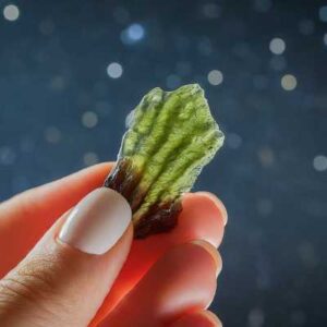A photo of a Moldavite crystal held against the backdrop of a starry night sky, with swirling galaxies visible.