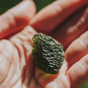 A close-up photo of a raw Moldavite crystal resting in an open palm, sunlight catching its textured green surface.
